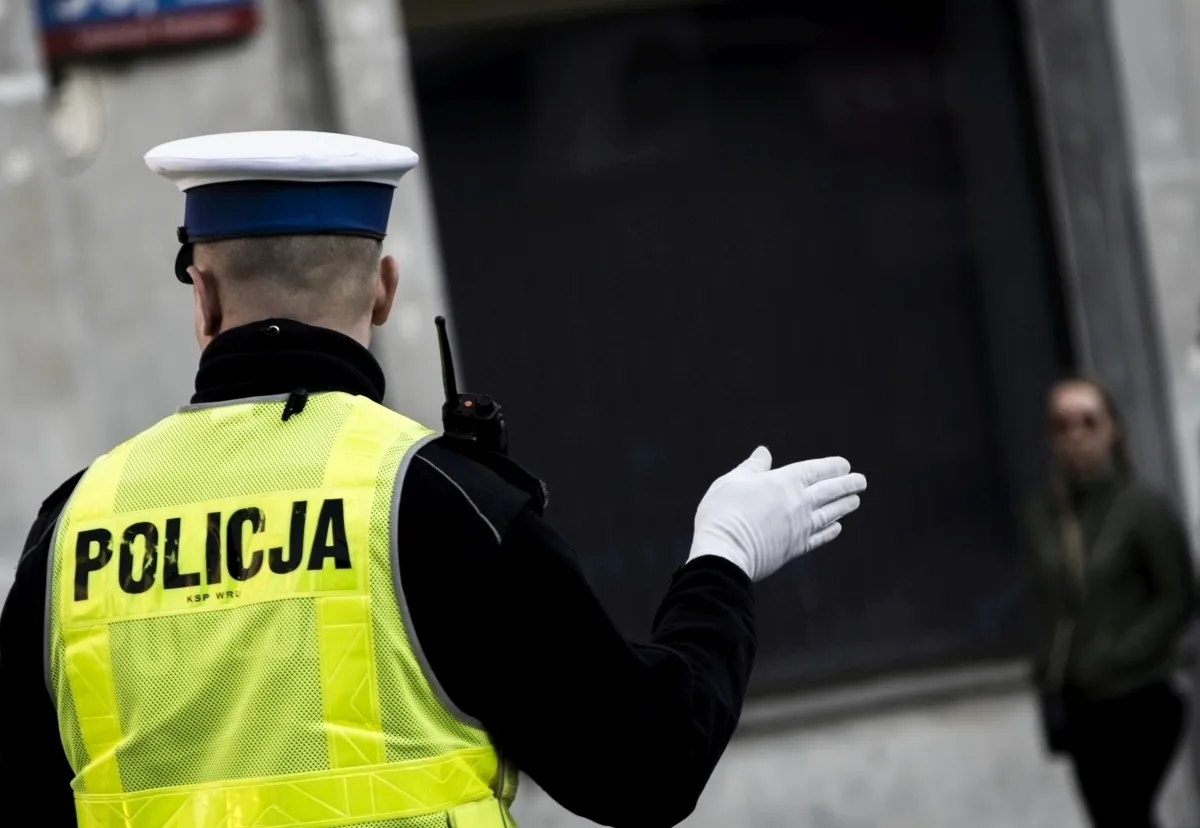 Police officer in a yellow POLICJA vest gesturing with a white glove toward a blurred woman